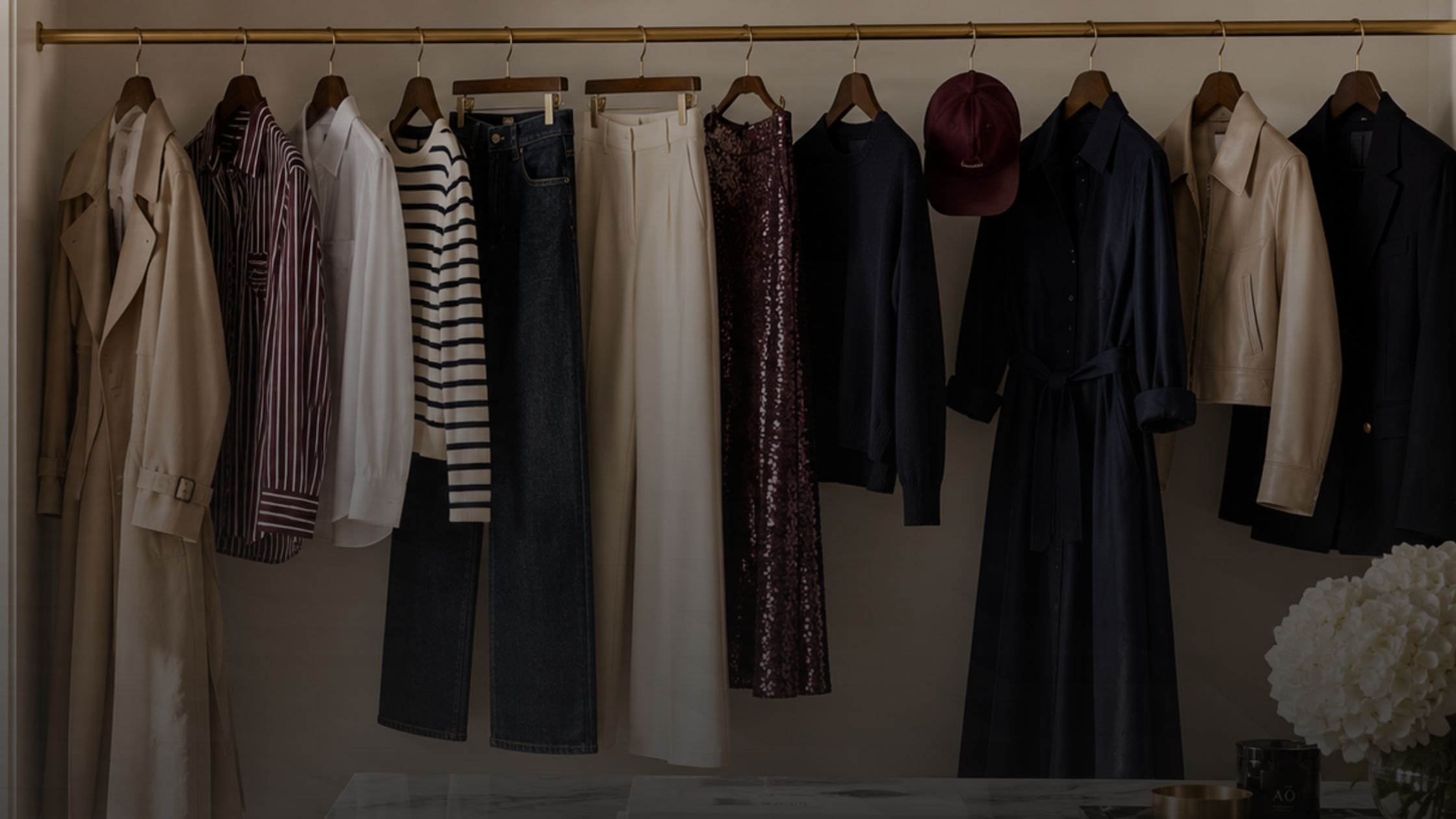 A curated wardrobe on a brass rail: trench, shirts, knitwear, denim, sequin skirt and tailoring in cream, navy and black.
