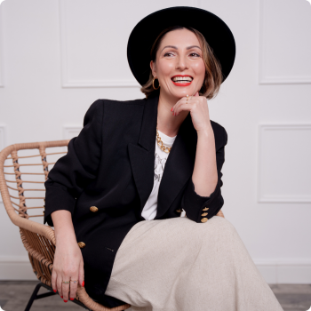 Roberta Lee - The Sustainable Stylist sits in a chair laughing. She wears a hat and black blazer with cream skirt .